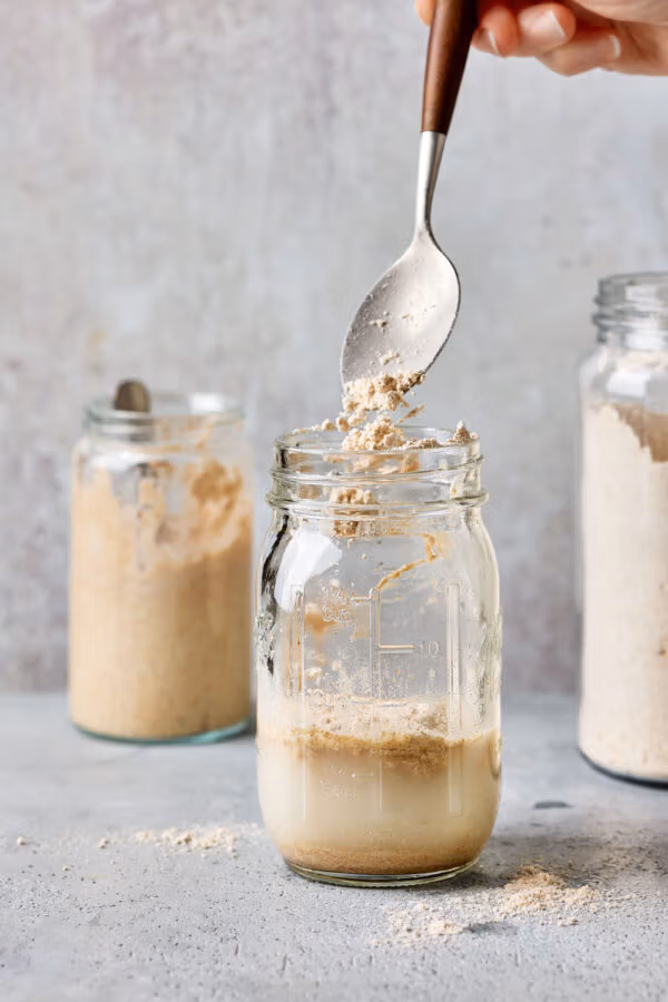 Mehl wird in eine Einmachglas mit Wasser und Sauerteiganstellgut gegeben, um den Sauerteig zu füttern.