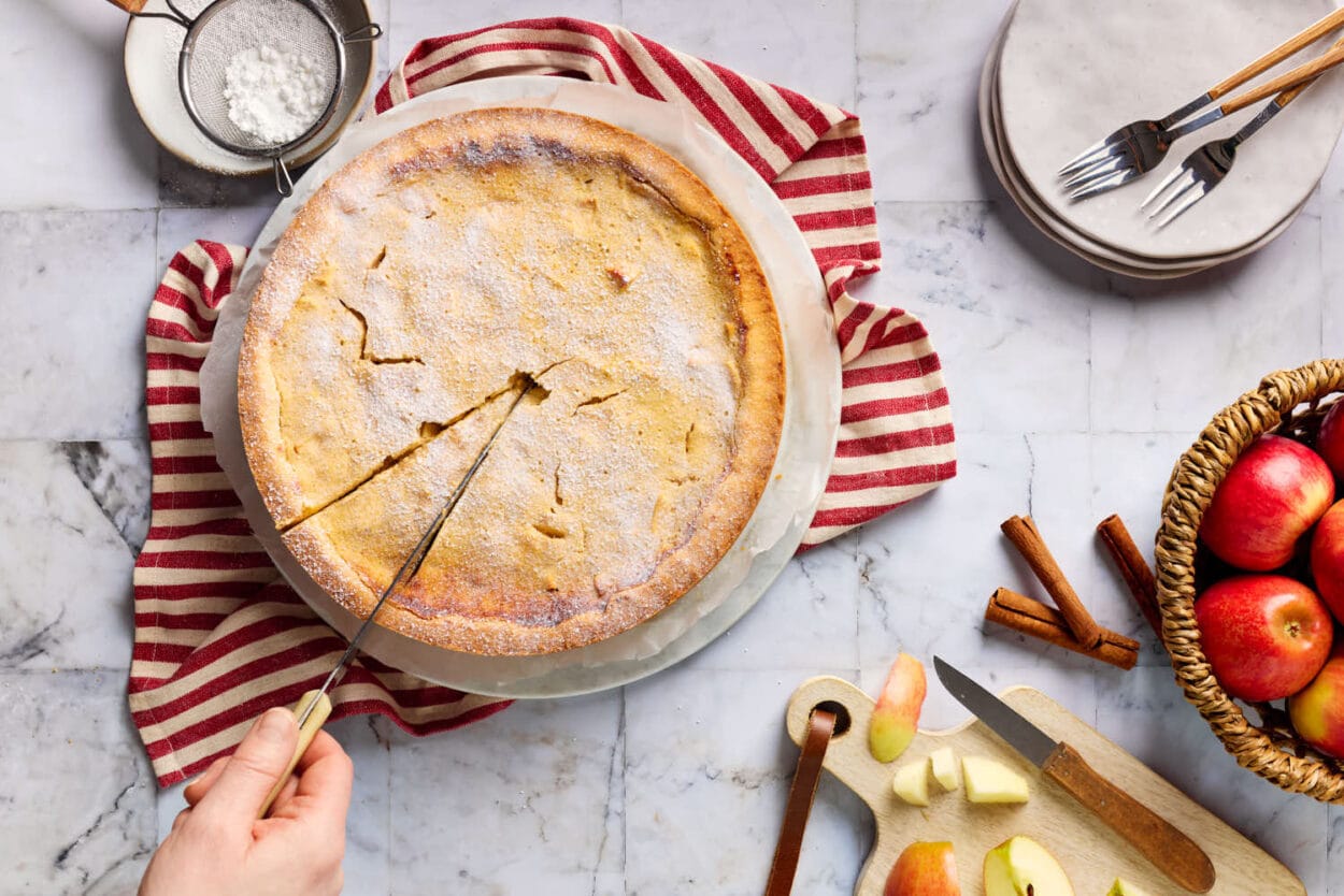 Saftiger Apfel-Quark-Kuchen mit zartem Mürbeteigboden in der Draufsicht, der angeschnitten wird. Drumherum sind mit Äpfeln, ein Sieb mit Puderzucker und Kuchenteller mit Kuchengabeln zu sehen.