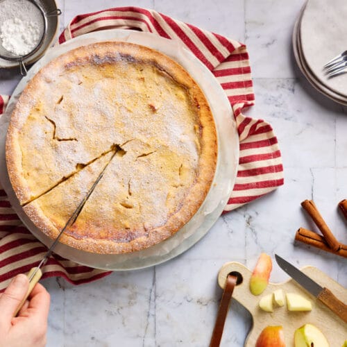Saftiger Apfel-Quark-Kuchen mit zartem Mürbeteigboden in der Draufsicht, der angeschnitten wird. Drumherum sind mit Äpfeln, ein Sieb mit Puderzucker und Kuchenteller mit Kuchengabeln zu sehen.