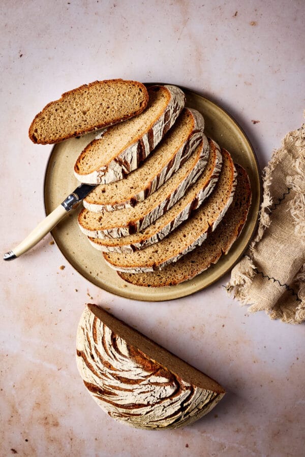 Roggen-Dinkel-Brot mit Sauerteig, aufgeschnitten auf einem Keramikteller mit einem Leinentuch und einem Brotmesser.