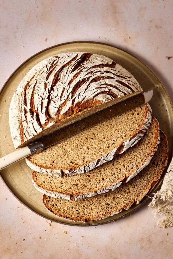 Roggen-Dinkel-Brot mit Sauerteig, aufgeschnitten auf einem Keramikteller mit einem Leinentuch und einem Brotmesser.