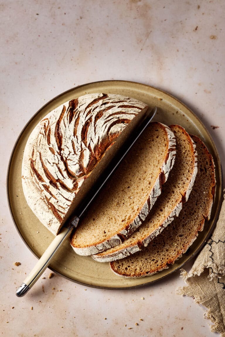 Roggen-Dinkel-Brot mit Sauerteig, aufgeschnitten auf einem Keramikteller mit einem Leinentuch und einem Brotmesser.