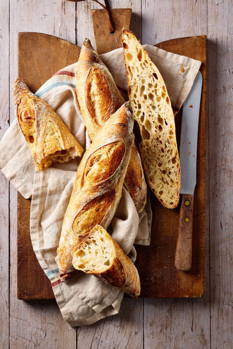 Baguette mit Sauerteig und lockerer Krume auf einem französischen Holzbrett mit einem Leinentuch und Brotmesser daneben.