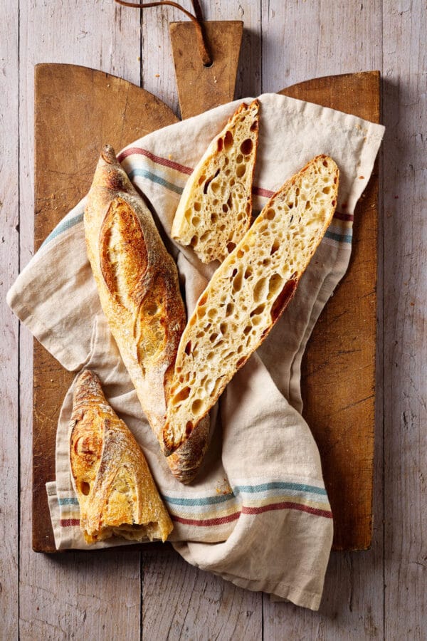 Baguette mit Sauerteig und lockerer Krume auf einem französischen Holzbrett mit einem Leinentuch und Brotmesser daneben.