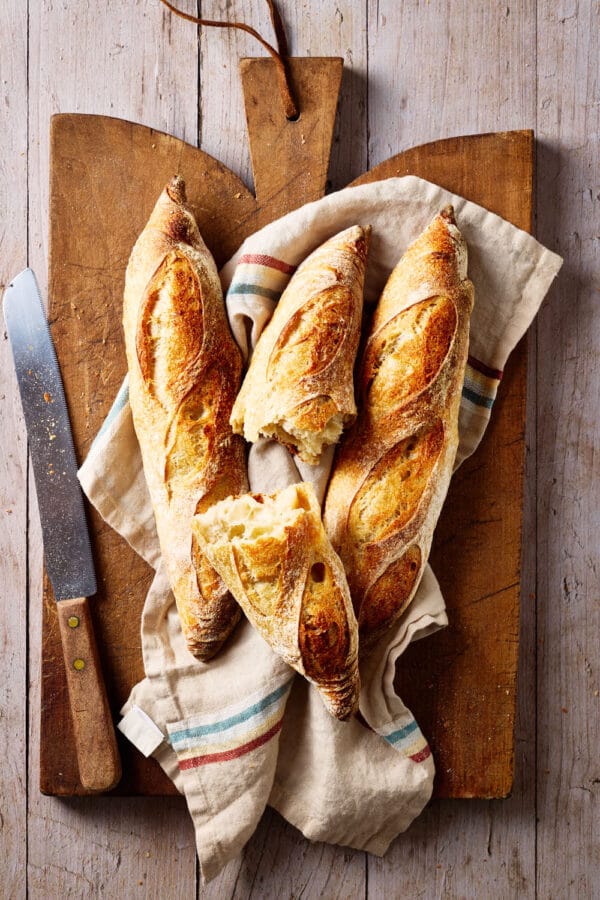 Baguette mit Sauerteig auf einem französischen Holzbrett mit einem Leinentuch und Brotmesser daneben.