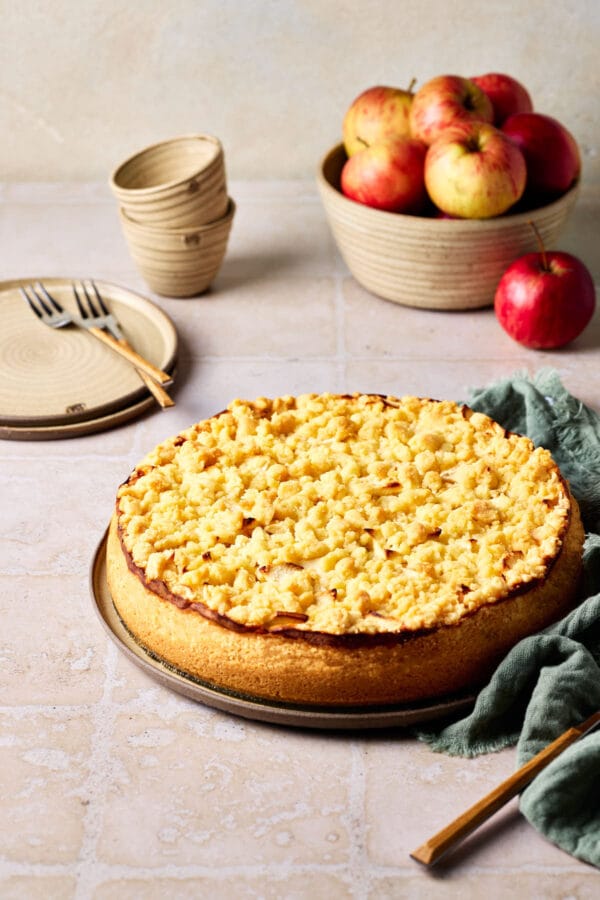 Saftiger Apfelkuchen mit Quark und Streusel auf einem Teller, mit Äpfeln im Hintergrund.