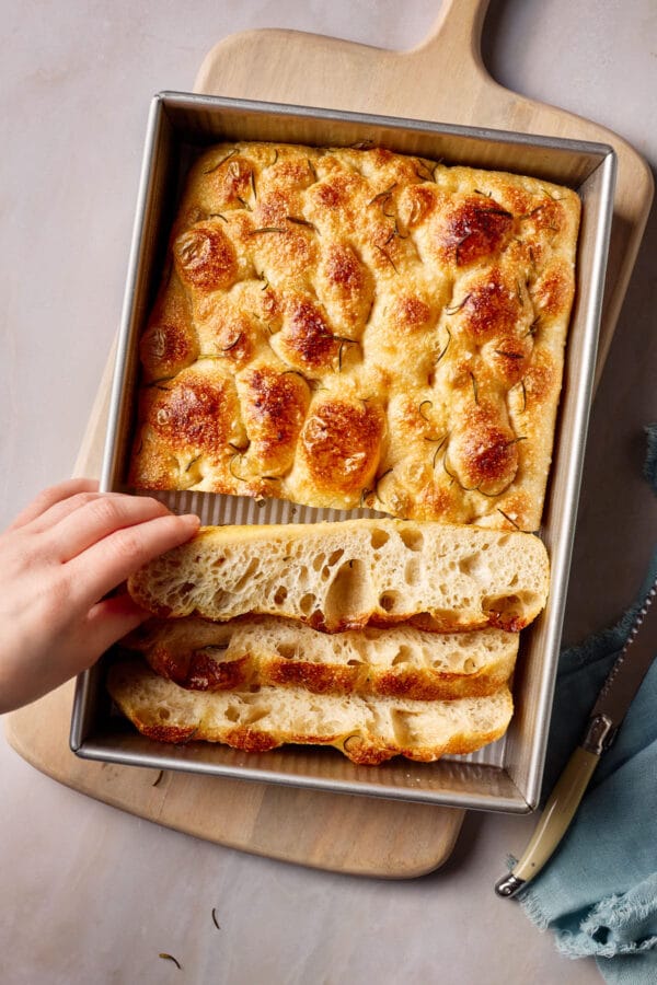 Luftige, angeschnittene Focaccia mit Sauerteig in eine Backform, ein Stück Focaccia wird von einer Hand herausgenommen.