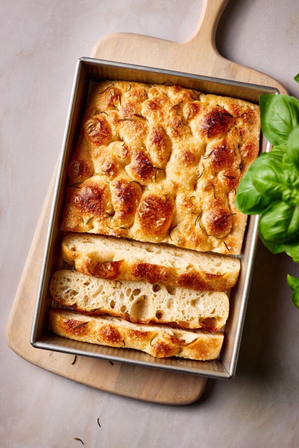 Luftige, angeschnittene Focaccia mit Sauerteig in eine Backform mit frischem Basilikum daneben.