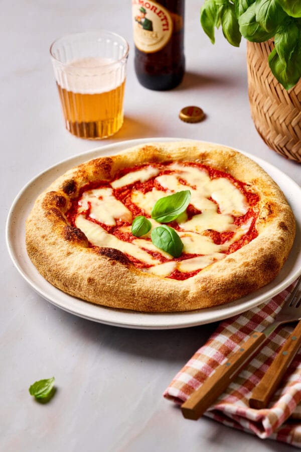 Frisch gebackene Sauerteig-Pizza auf einem Teller, mit einer karierten Serviette daneben und einem Glas Bier und Basilikum im Hintergrund.