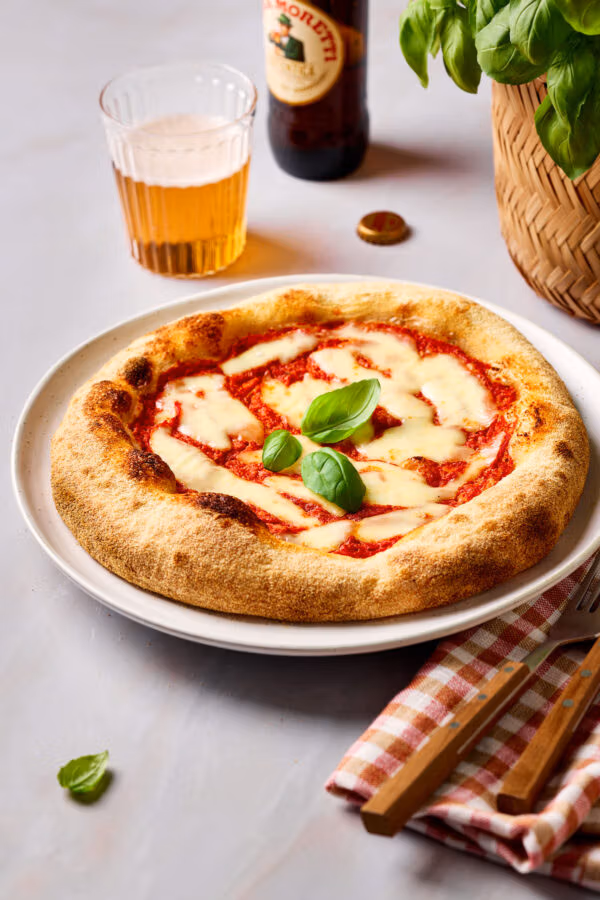 Frisch gebackene Sauerteig-Pizza auf einem Teller, mit einer karierten Serviette daneben und einem Glas Bier und Basilikum im Hintergrund.