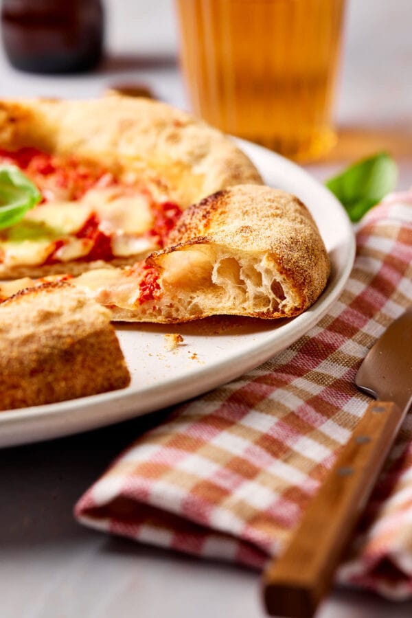 Frisch gebackene Sauerteig-Pizza auf einem Teller, mit einer karierten Serviette daneben und einem Glas Bier im Hintergrund.