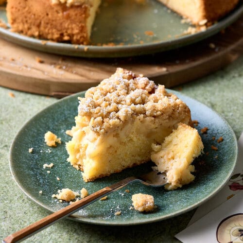 Ein Stück Apfel-Streuselkuchen auf einem Kuchenteller mit einer Gabel und zwei Postkarten daneben.