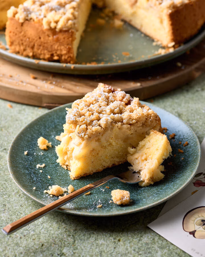 Ein Stück Apfel-Streuselkuchen auf einem Kuchenteller mit einer Gabel und zwei Postkarten daneben.