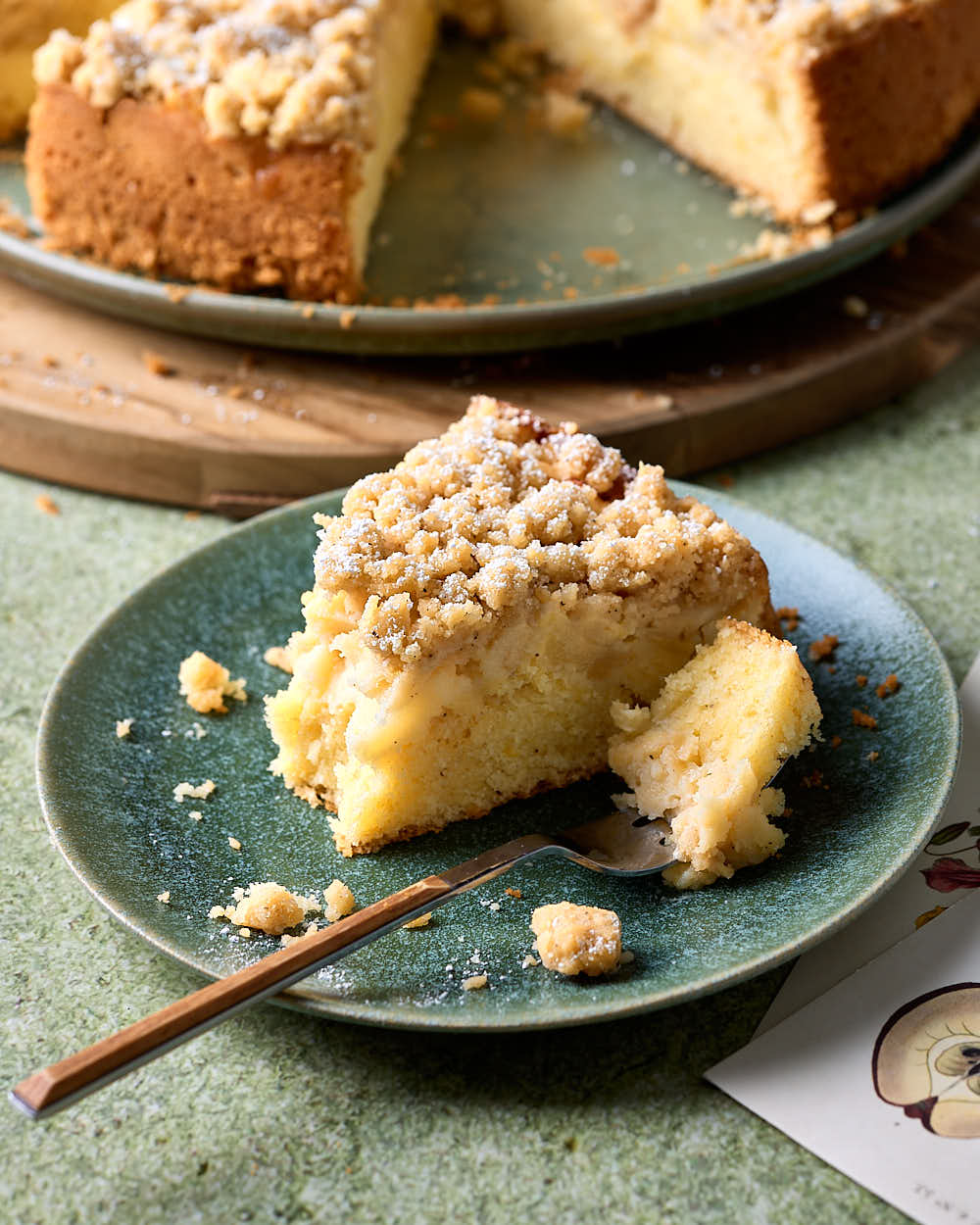 Ein Stück Apfel-Streuselkuchen auf einem Kuchenteller mit einer Gabel und zwei Postkarten daneben.