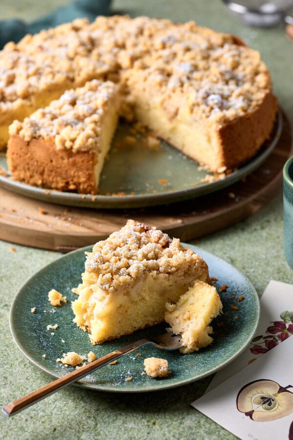 Ein Stück Apfel-Streuselkuchen auf einem Kuchenteller mit einer Gabel und zwei Postkarten daneben.