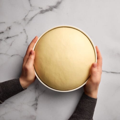 Hefeteig in einer Keramikschüssel, die von zwei Händen gehalten wird.