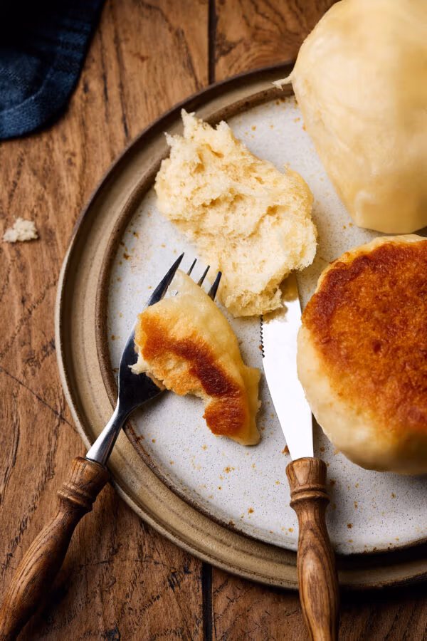 Dampfnudeln mit Salzkruste auf einem Teller mit lockerer Krume und Holzbesteck daneben.