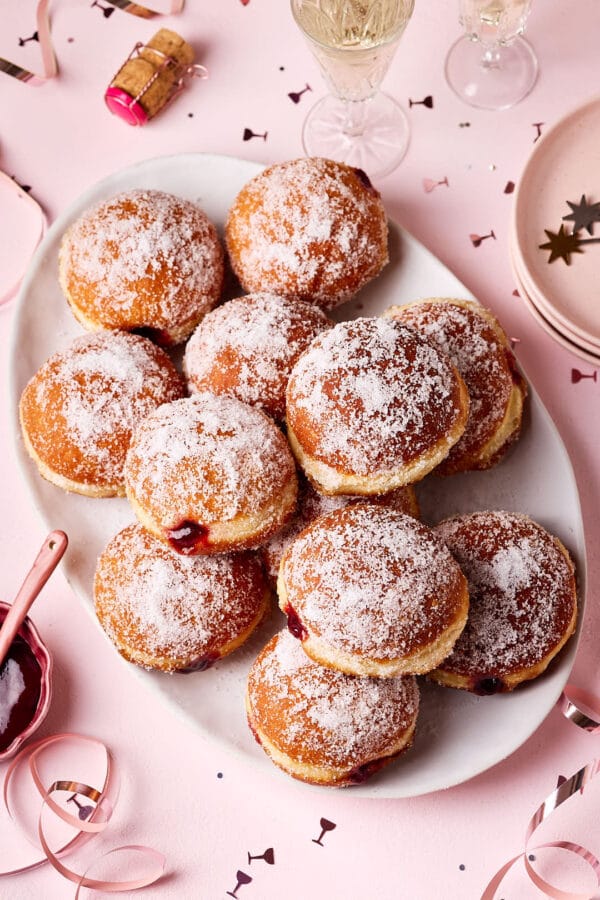 Frisch gebackene Berliner (Krapfen) aus Hefeteig auf einer Servierplatte m,t Silvesterdeko und Sektgläsern im Hintergrund.