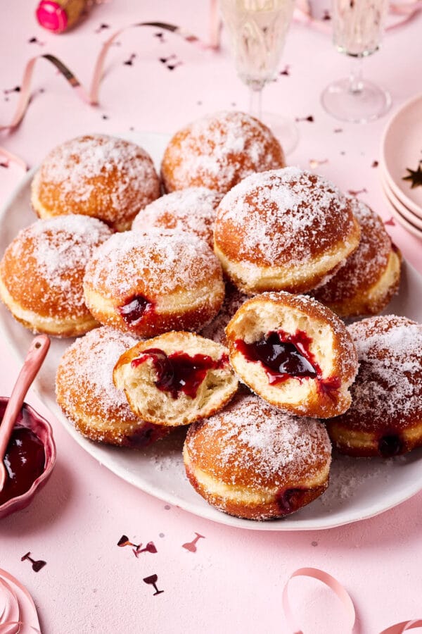 Frisch gebackene Berliner (Krapfen) aus Hefeteig, mit lockerer Krume und gefüllt mit Konfitüre, auf einer Servierplatte mit Silvesterdeko und Sektgläsern im Hintergrund.