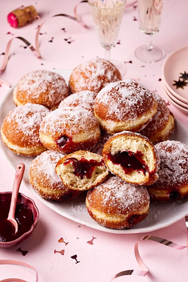 Frisch gebackene Berliner (Krapfen) aus Hefeteig, mit lockerer Krume und gefüllt mit Konfitüre, auf einer Servierplatte mit Silvesterdeko und Sektgläsern im Hintergrund. 