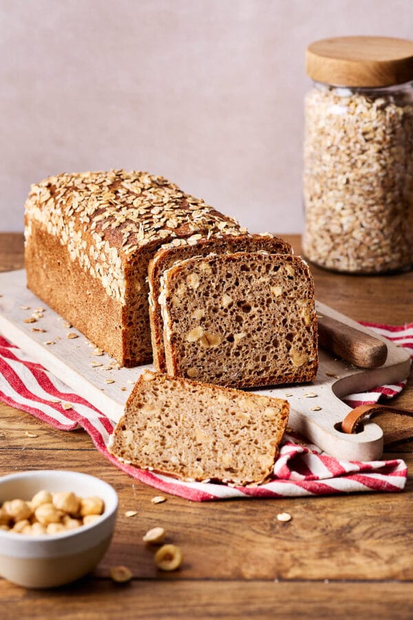 Einfaches Nussbrot mit Vollkornmehl aus der Kastenform auf einem Holzbrett, mit einem Glas Haferflocken im Hintergrund und einer Schale Haselnüsse im Vordergund.
