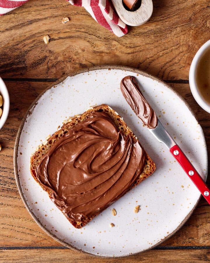 Eine Scheibe Nussbrot mit Nuss-Nougat-Creme auf einem Teller mit einem Messer daneben.