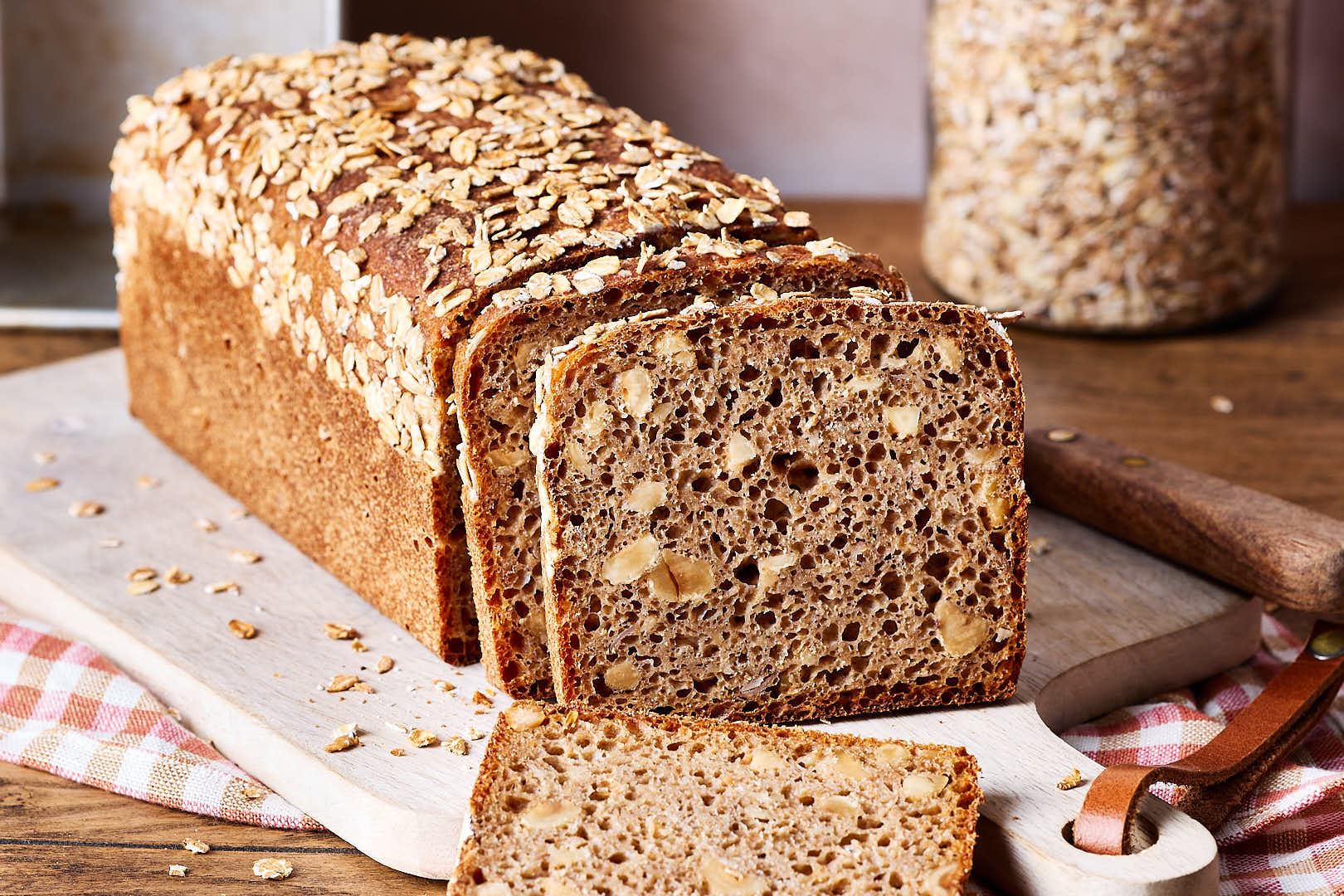 Einfaches Nussbrot mit Vollkornmehl aus der Kastenform auf einem Holzbrett, mit einem Glas Haferflocken im Hintergrund und einer Schale Haselnüsse im Vordergund.
