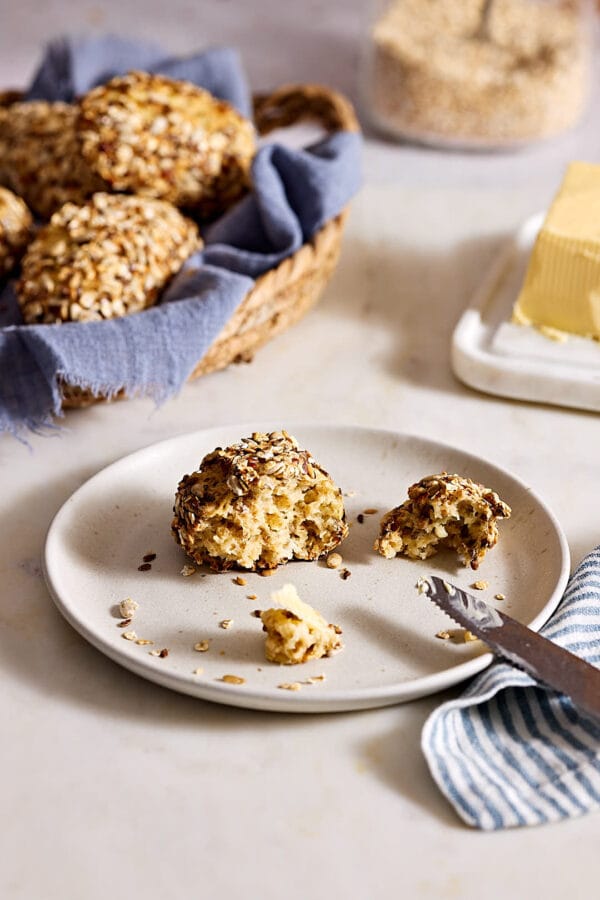Eiweißbrötchen mit Quark, mit lockerer Krume, auf einem Teller mit etwas Butter sowie einem Korb mit Brötchen und Butter im Hintergrund.