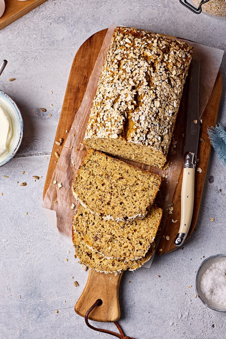 Schnelles Eiweißbrot mit Quark, aufgeschnitten auf einem Holzbrett mit Eiern, Haferflocken, Leinsamen und Quark im Hintergrund und einem Schälchen Salz im Vordergrund.