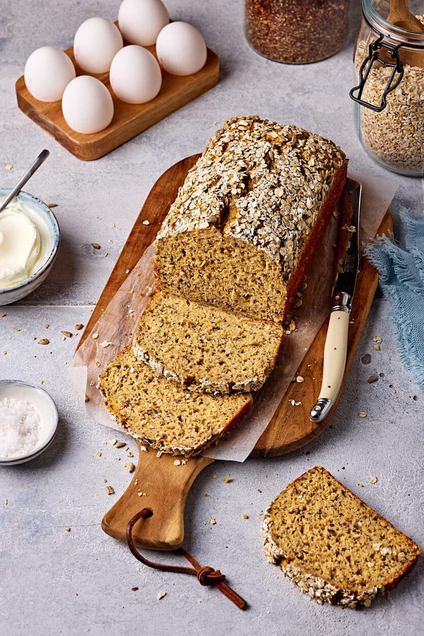 Schnelles Eiweißbrot mit Quark, aufgeschnitten auf einem Holzbrett mit Eiern, Haferflocken, Leinsamen und Quark im Hintergrund und einem Schälchen Salz im Vordergrund.