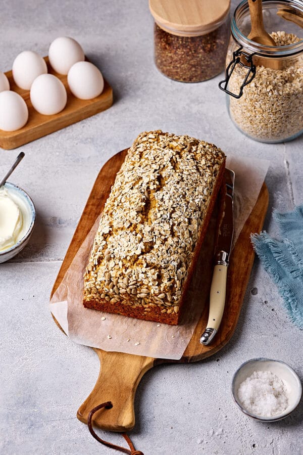 Schnelles Eiweißbrot mit Quark auf einem Holzbrett mit Eiern, Haferflocken, Leinsamen und Quark im Hintergrund und einem Schälchen Salz im Vordergrund.