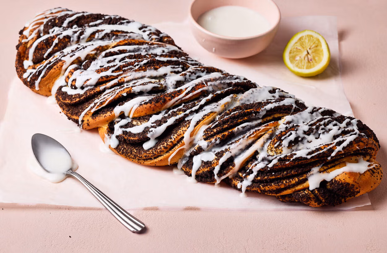 Saftiger Mohnzopf mit Zuckerguss auf einem Stück Backpapier mit einem Löffel im Vordergrund.