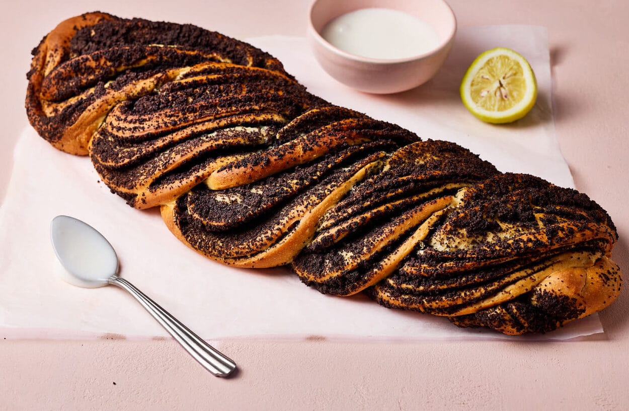 Saftiger Mohnzopf auf einem Stück Backpapier mit einem Löffel im Vordergrund und einer Schale Zuckerguss und einer halben Zitrone im Hintergrund.