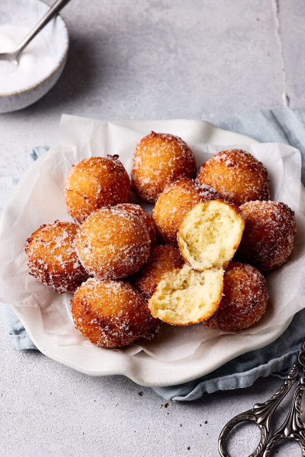 Luftige Quarkbällchen mit Zuckerkruste auf einem Teller mit einer Schale mit Zucker im Hintergrund. 