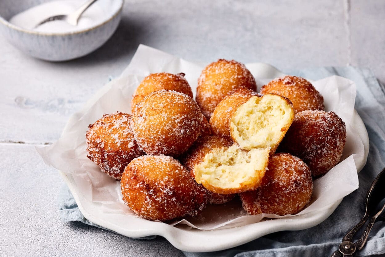 Luftige Quarkbällchen mit Zuckerkruste auf einem Teller mit einer Schale mit Zucker im Hintergrund.