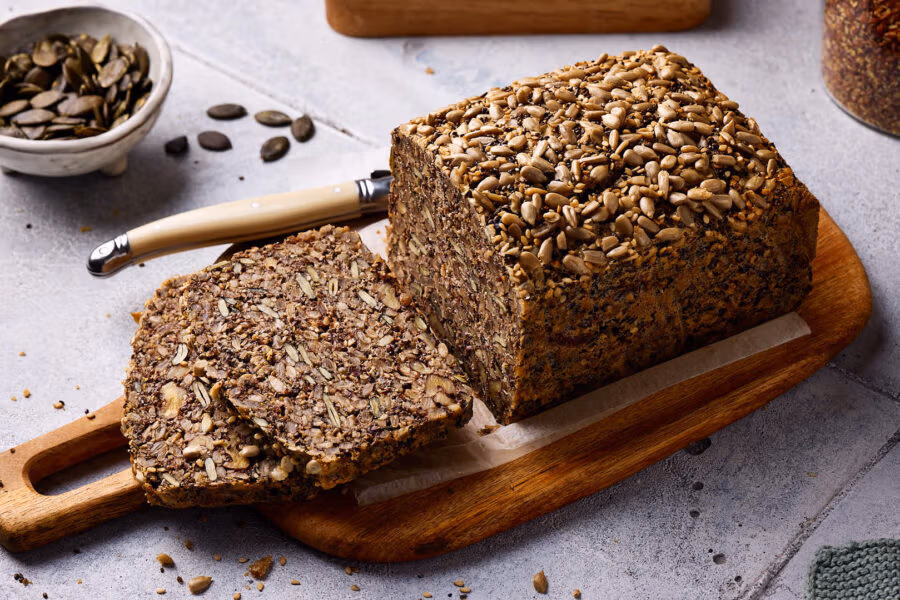 Saatenbrot ohne Mehl aufgeschnitten auf einem Holzbrett mit Zutaten im Hintergrund.