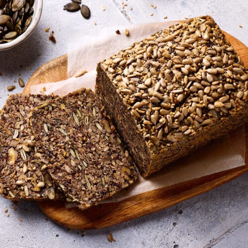 Saatenbrot ohne Mehl aufgeschnitten auf einem Holzbrett mit Zutaten im Hintergrund.