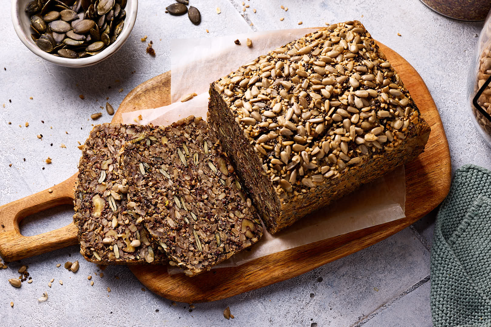 Saatenbrot ohne Mehl aufgeschnitten auf einem Holzbrett mit Zutaten im Hintergrund.