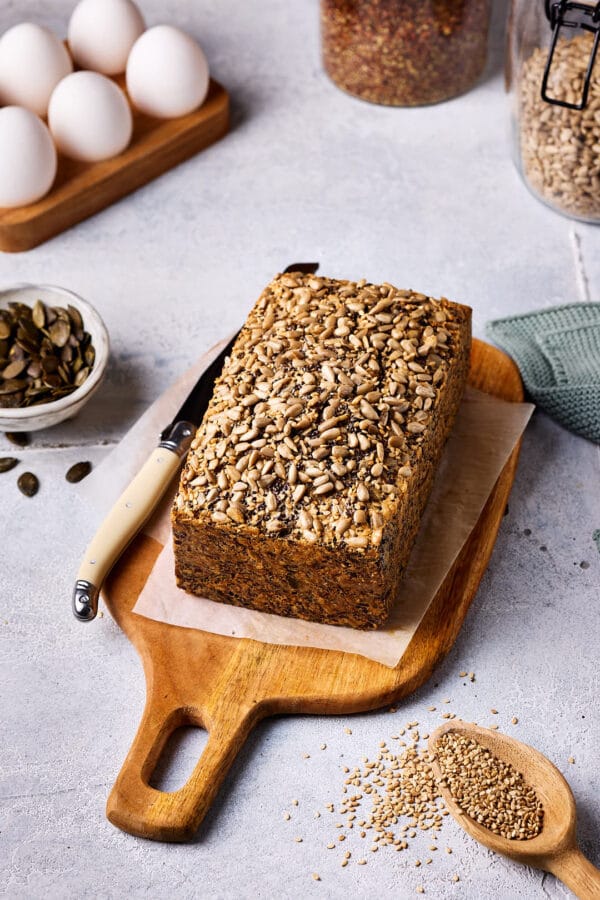 Saatenbrot ohne Mehl auf einem Holzbrett mit einem Messer daneben und weiteren Zutaten im Hintergrund.