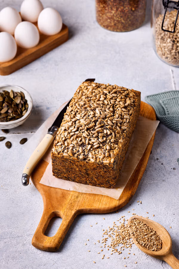 Saatenbrot ohne Mehl auf einem Holzbrett mit einem Messer daneben und weiteren Zutaten im Hintergrund.