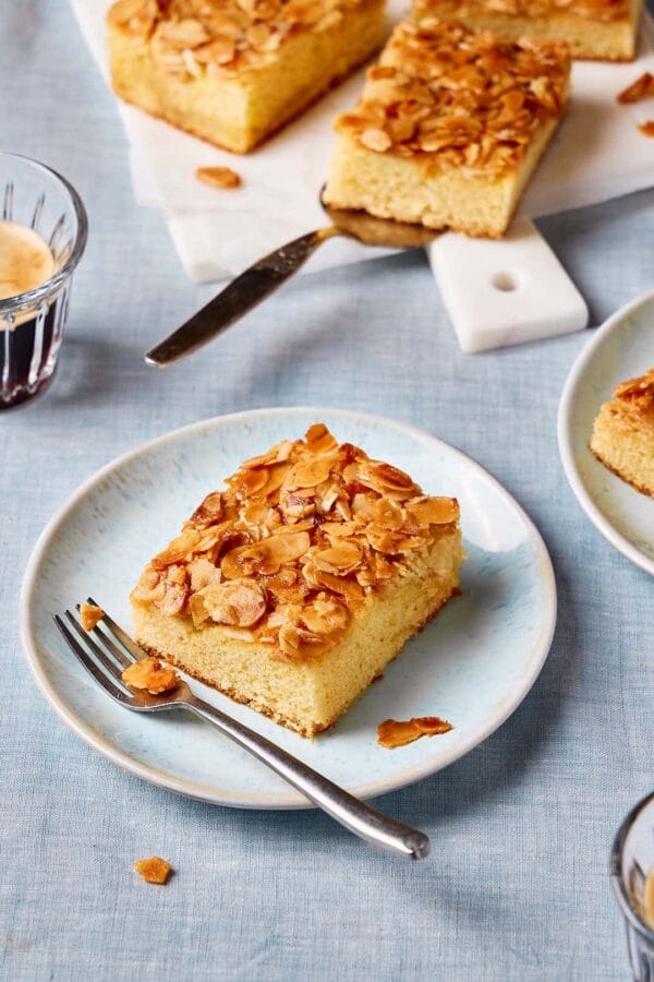 Saftiger Butterkuchen ohne Hefe vom Blech auf einem Teller mit Kuchenstücken und Kaffeetassen im Hintergrund. 