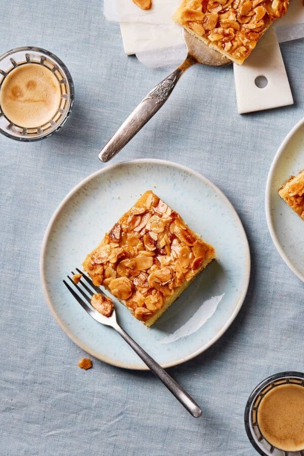 Saftiger Butterkuchen ohne Hefe vom Blech auf einem Teller mit Kuchenstücken und Kaffeetassen im Hintergrund. 
