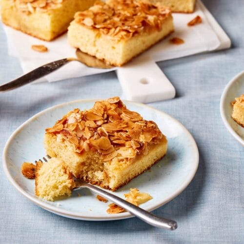 Saftiger Butterkuchen ohne Hefe vom Blech auf einem Teller mit Kuchenstücken und Kaffeetassen im Hintergrund.