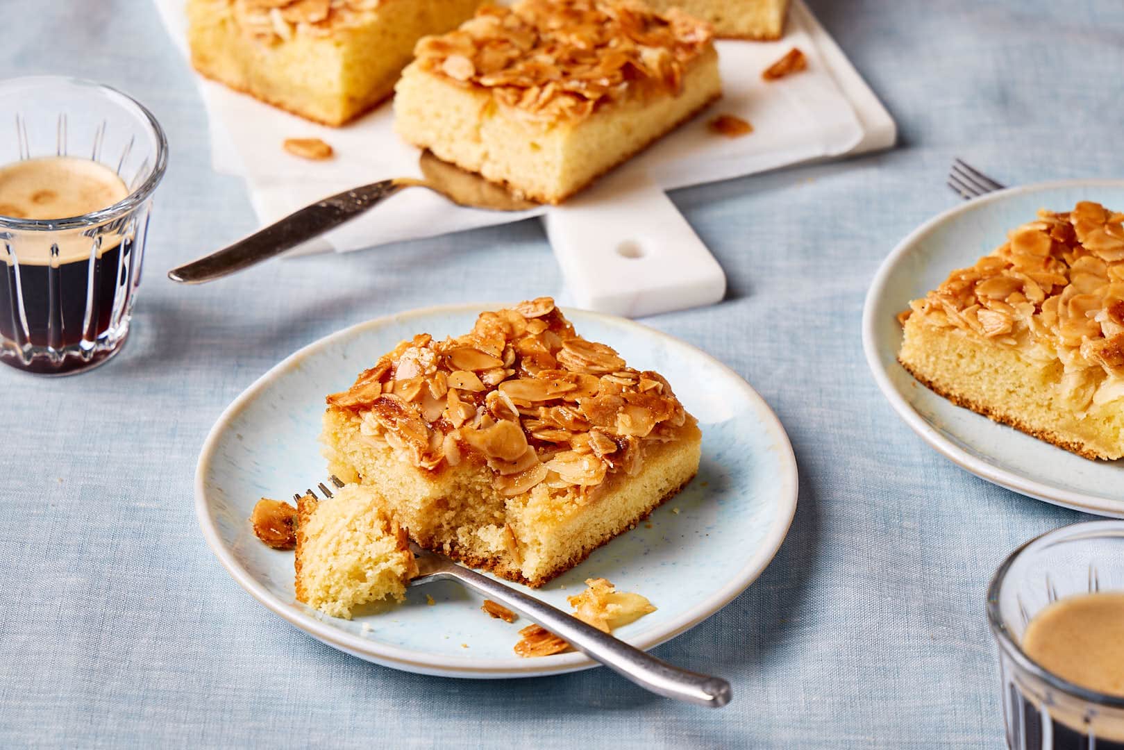 Saftiger Butterkuchen ohne Hefe vom Blech auf einem Teller mit Kuchenstücken und Kaffeetassen im Hintergrund.