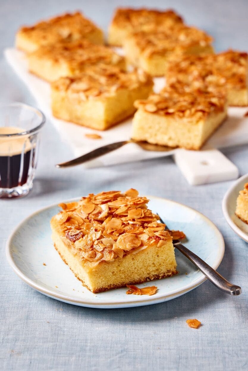 Saftiger Butterkuchen ohne Hefe vom Blech auf einem Teller mit Kuchenstücken und Kaffeetassen im Hintergrund.
