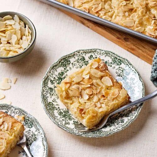 Omas Butterluchen mit Sahneguss und Mandel-Zucker-Topping auf Tellern mit einem Schälchen Mandeln im Hintergrund.