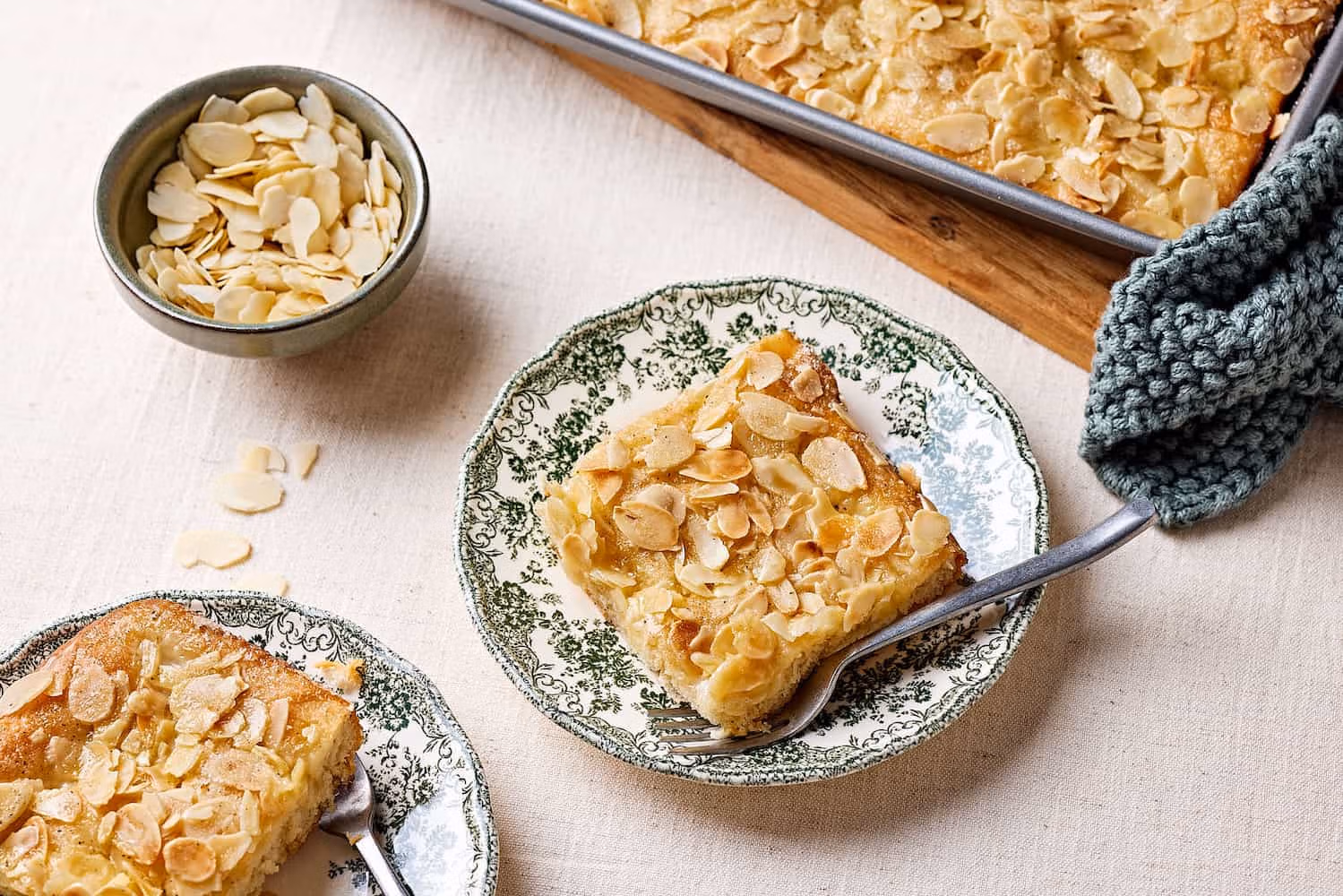 Omas Butterluchen mit Sahneguss und Mandel-Zucker-Topping auf Tellern mit einem Schälchen Mandeln im Hintergrund.