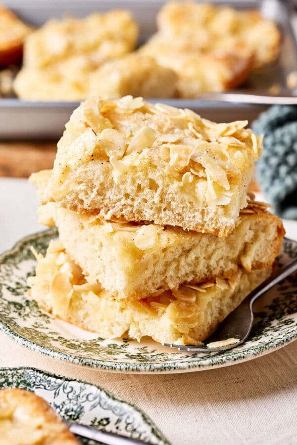 Omas Butterluchen mit Sahneguss und Mandel-Zucker-Topping auf Tellern mit einem Blech Kuchenstücke im Hintergrund.