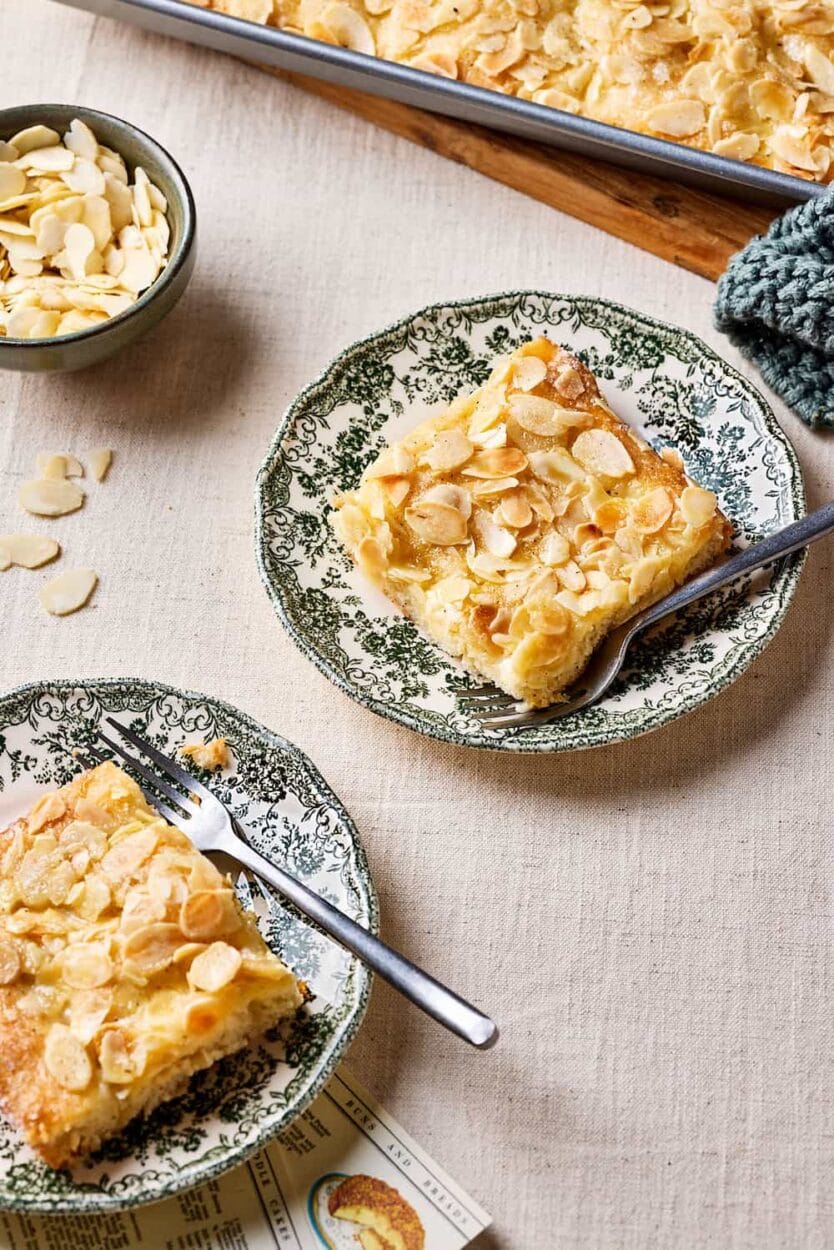 Omas Butterluchen mit Sahneguss und Mandel-Zucker-Topping auf Tellern mit einem Schälchen Mandeln im Hintergrund.