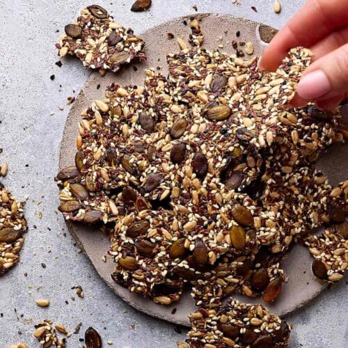 Saatencracker ohne Mehl auf einer Steingutplatte mit Körner-Crackern drumherum. Eine Hand greift nach einem Cracker.